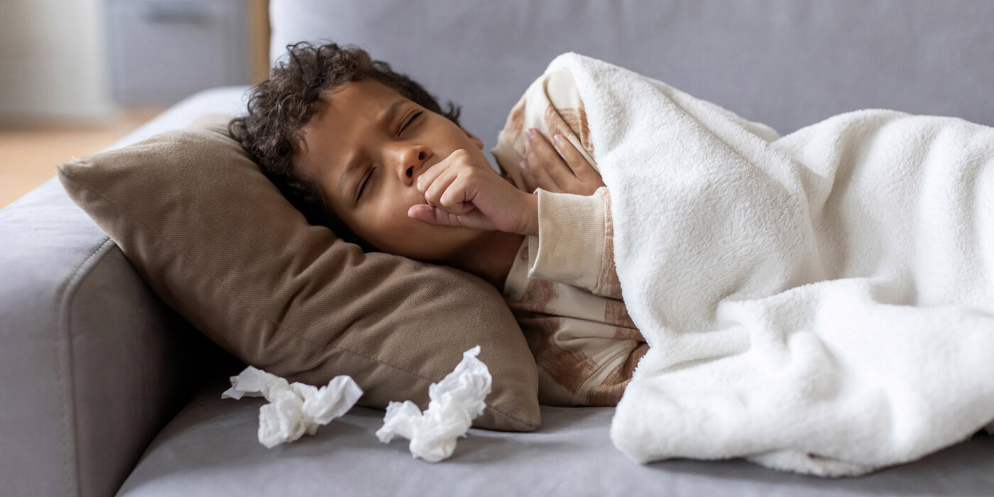 A young boy lies on a couch covered with used tissues and coughs
