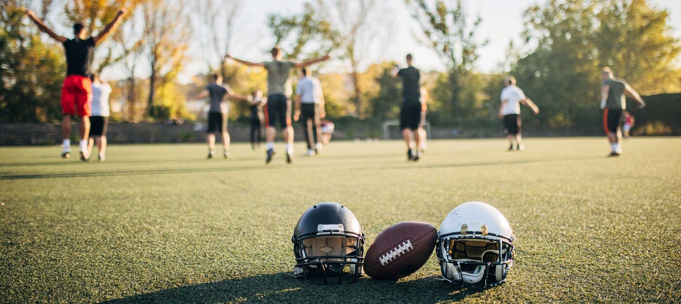 Two football helmets and a football sit int he foreground as a group of players cheers in the background.