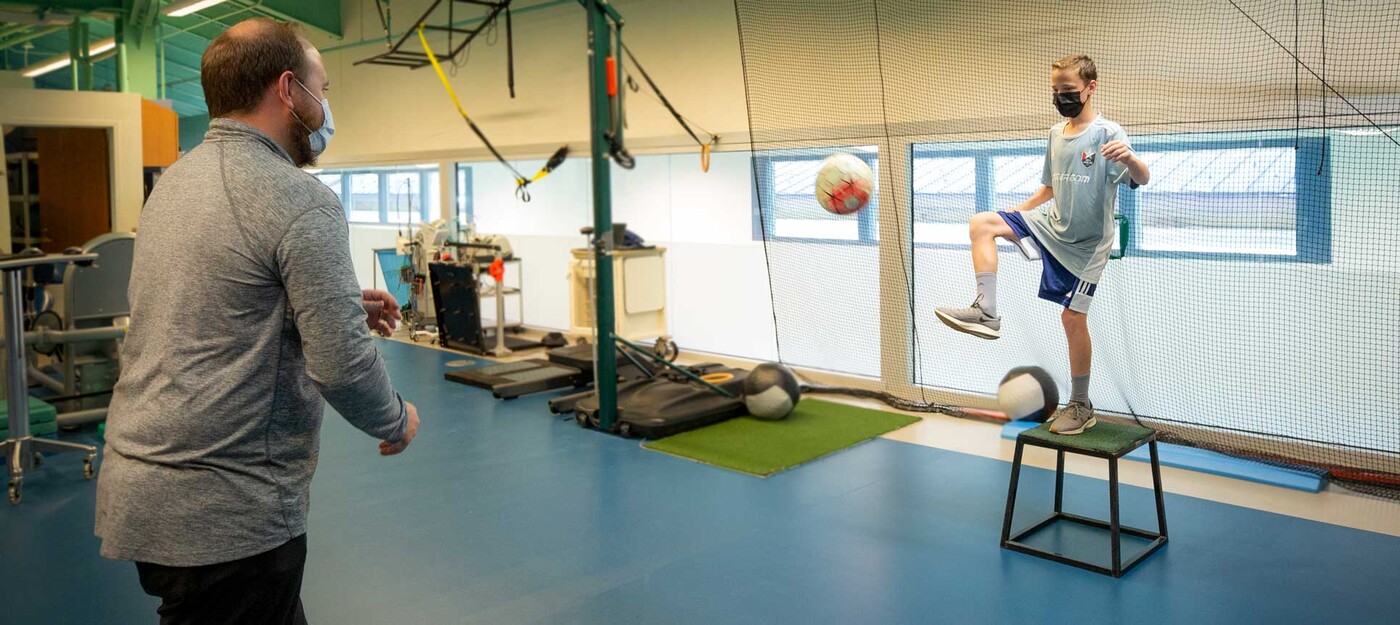 Mike Greeson works with physical therapist, Nicholas Mahaffey, as he continues to rehab his knee after an ACL repair. A physical therapist works with a young patient as he practices kicking a soccer ball while balanced on a raised platform.