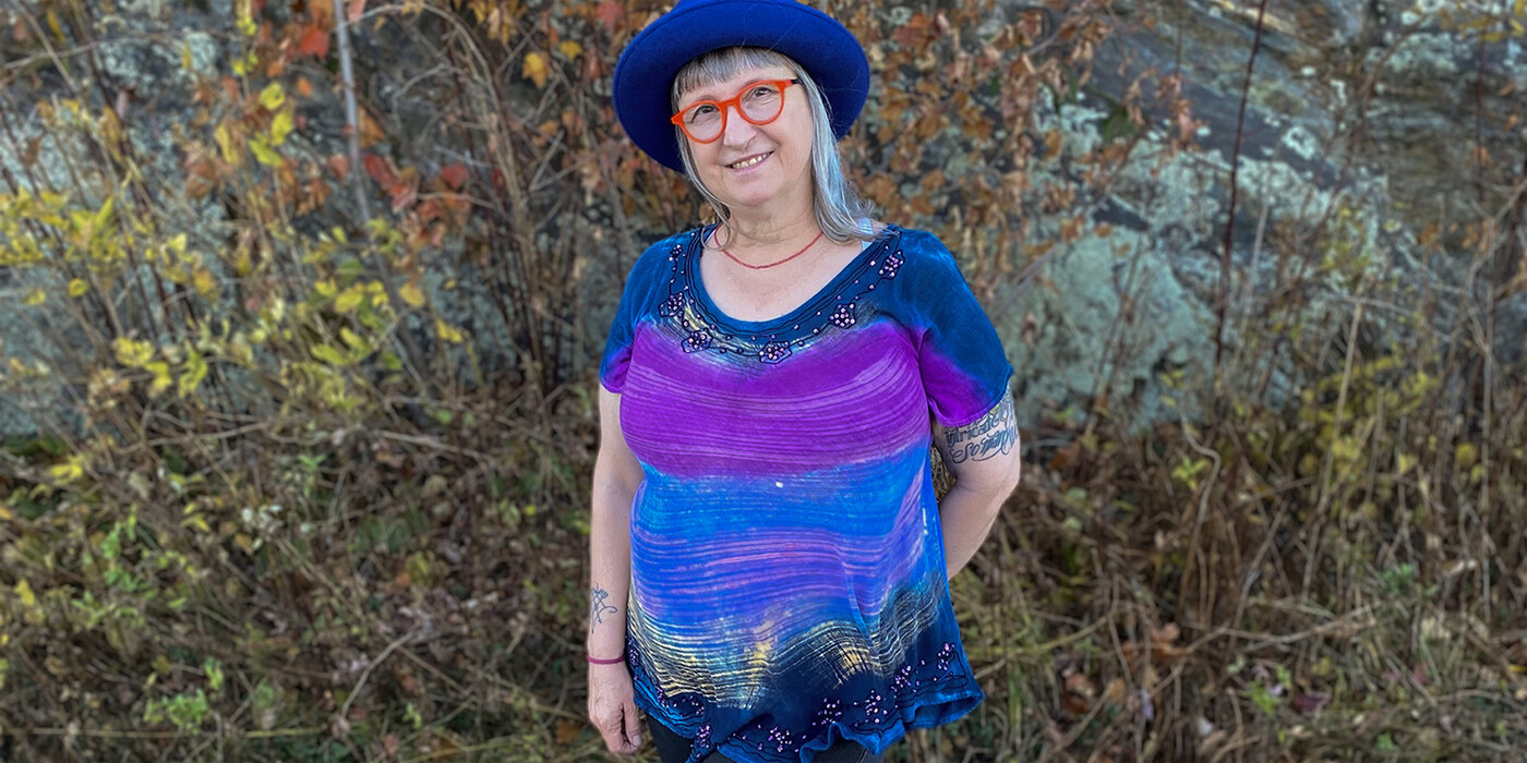 Beth Huber, 60, lives in Asheville, NC with her husband, children, and grandchildren. Beth Huber stands in front of some greenery. She's wearing a blue hat, orange glasses, and a blue and purple shirt.