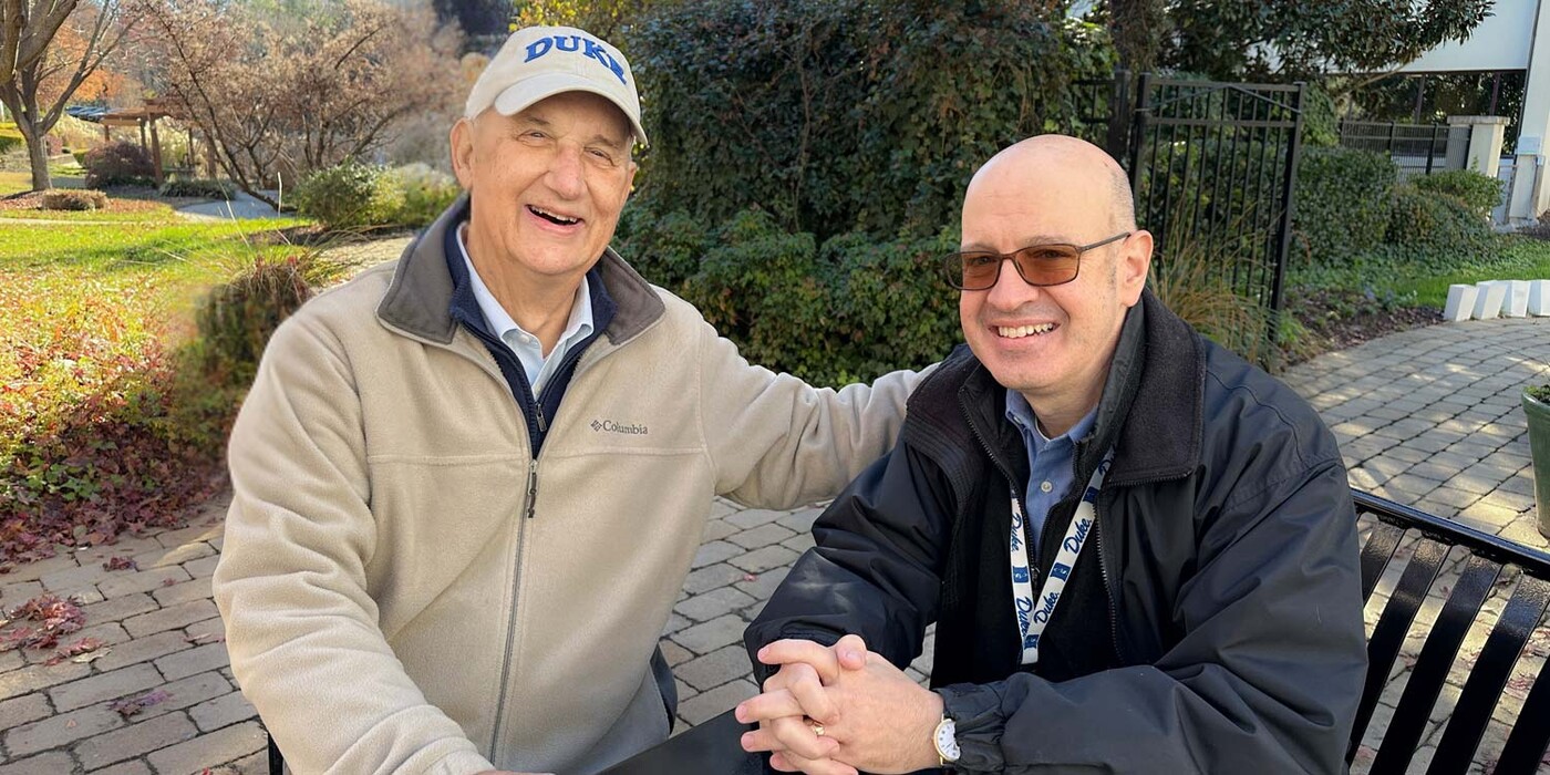 (Left to right): Art Krueger with Patrick Plumeri, MS, LFMT, a medical family therapist, at Duke Cancer Center Raleigh. Two men sit outside at a table, smiling at the camera